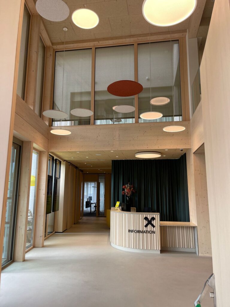 Zweigeschoßiges Foyer der Raiffeisenbank Gamlitz in Holzbauweise mit runden Pendelleuchten, Akustikelementen und Informationstheke